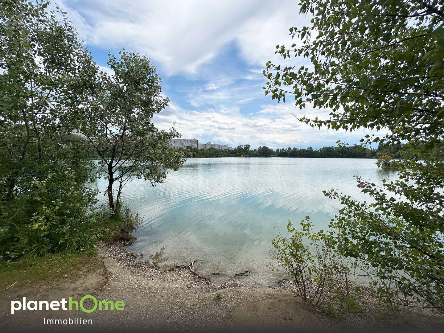 Malerischer Blick auf einen ruhigen See, umgeben von Bäumen unter einem bewölkten Himmel.