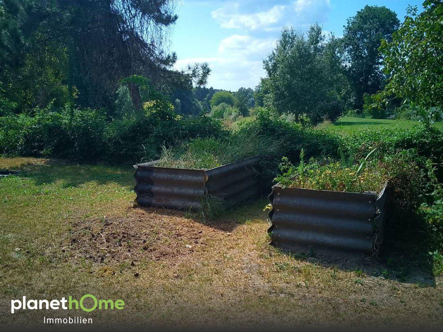 Gepflegter Gartenbereich mit Hochbeeten, umgeben von üppigem Grün und Bäumen unter blauem Himmel.