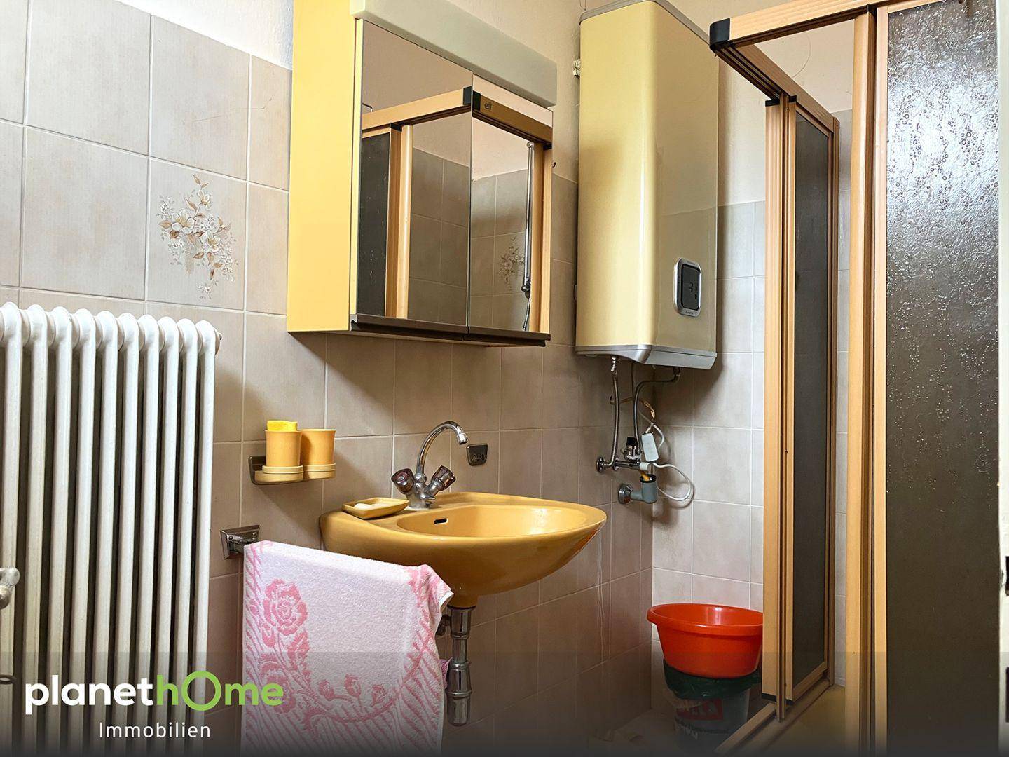 Badezimmer mit beigen Fliesen, einem Waschbecken, Dusche und einem Warmwasserboiler.