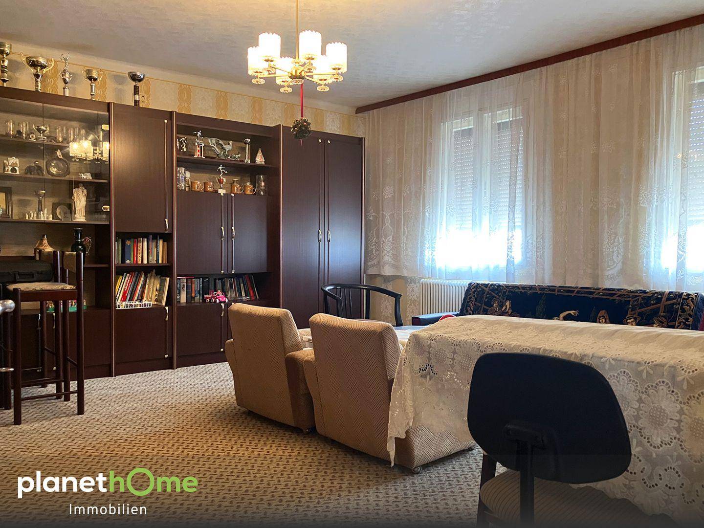 Geräumiges Wohnzimmer mit Teppichboden, einer großen Schrankwand und einem Kronleuchter.