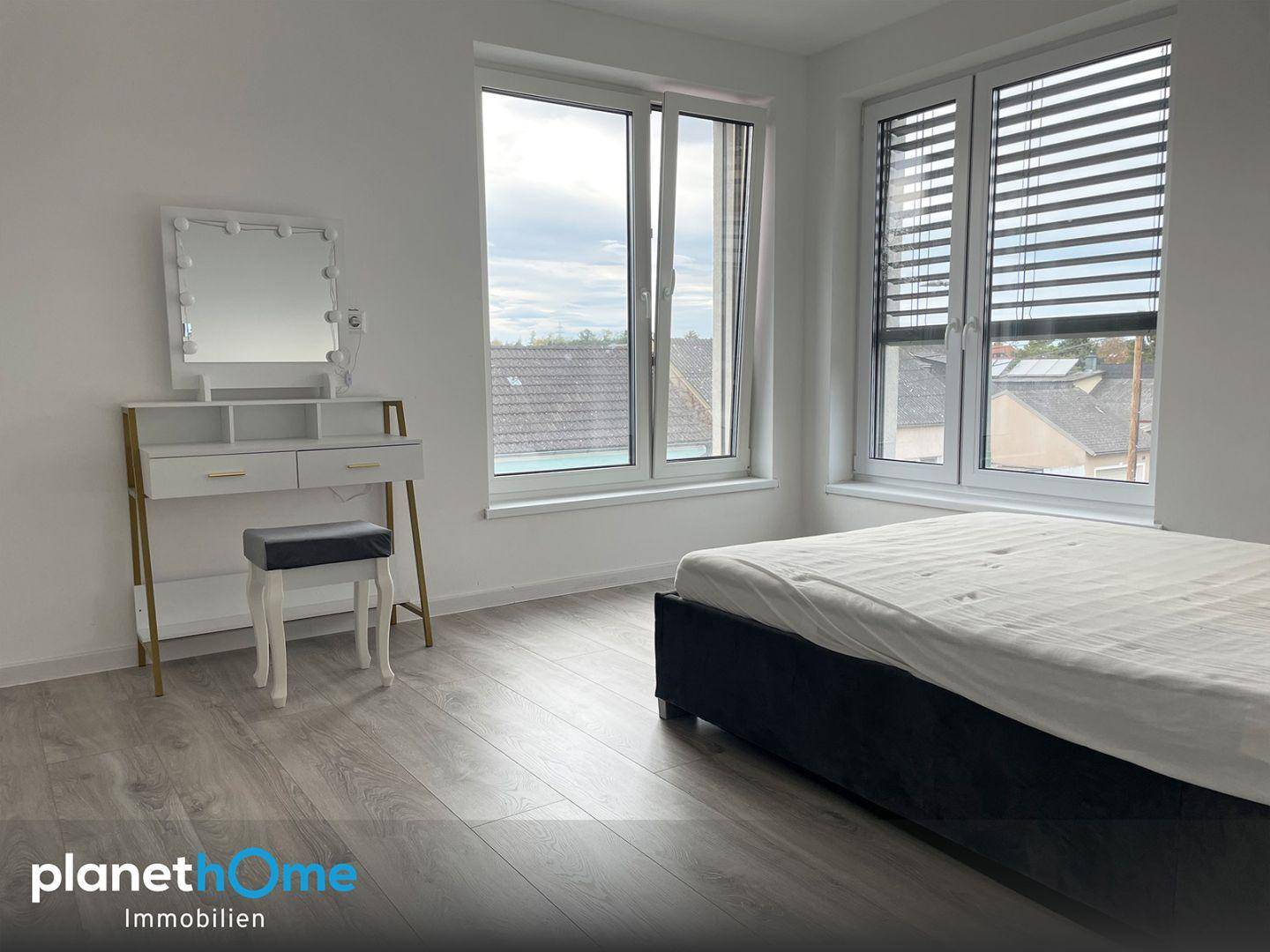 Helles Schlafzimmer mit großen Fenstern, Schminktisch und Laminatboden.
