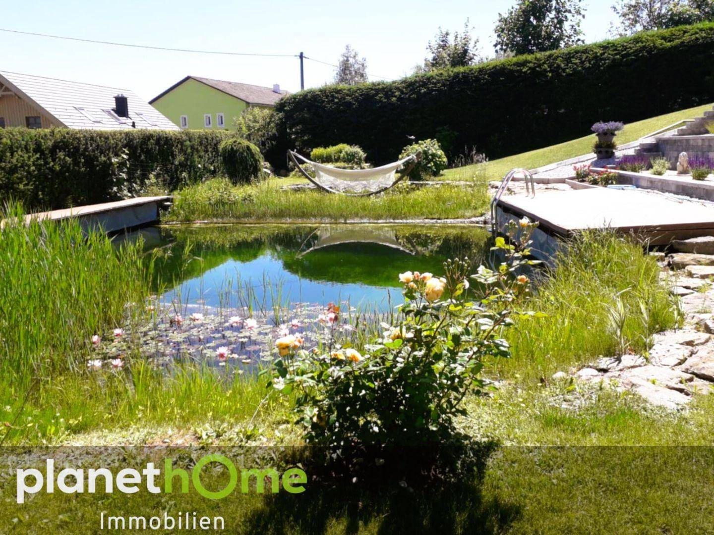 Idyllischer Naturteich mit Holzsteg, Hängematte und blühenden Rosen im Garten.