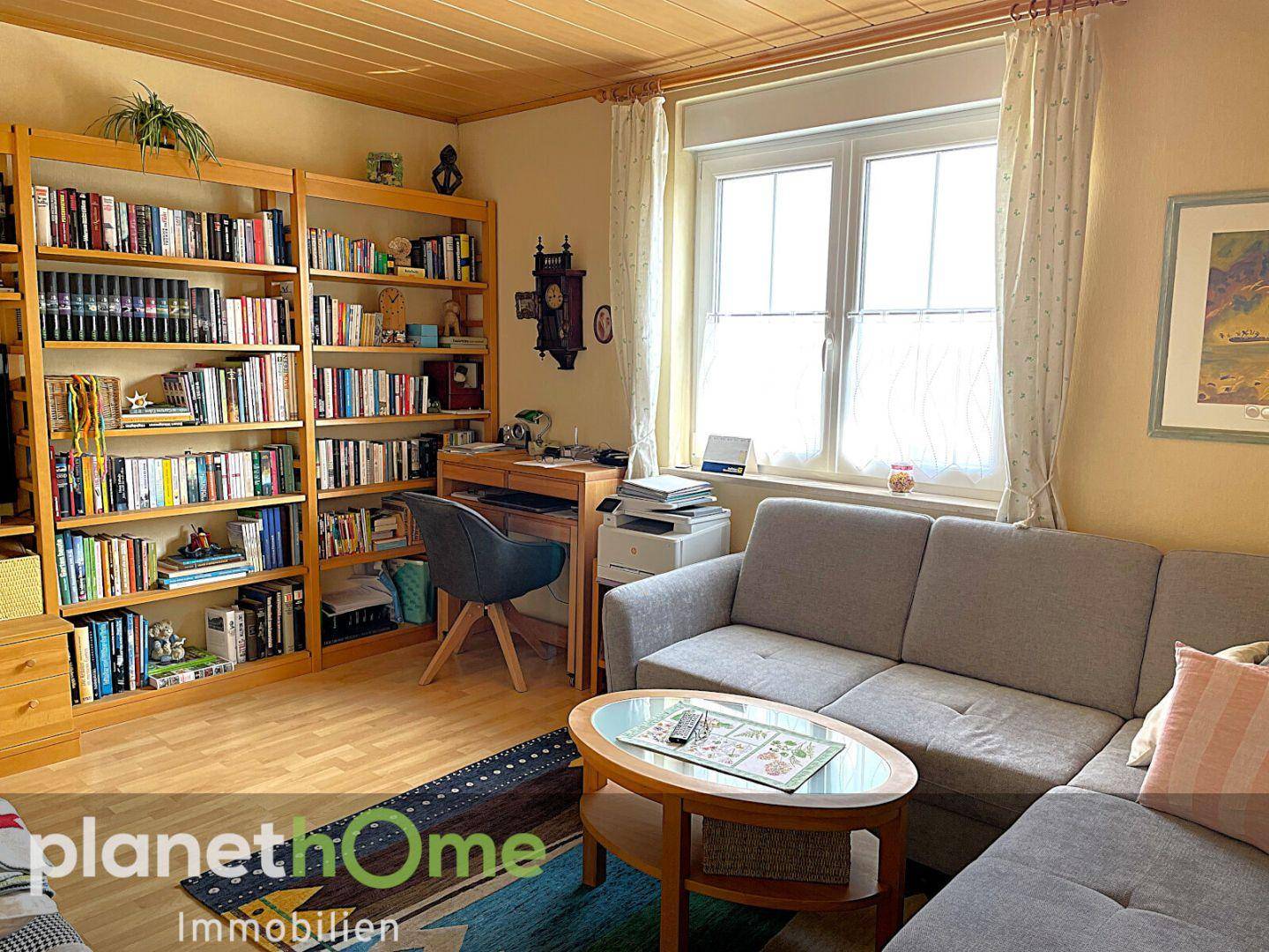 Heller Wohnbereich mit Sofa, Bücherregalen und einem Arbeitsplatz am Fenster.