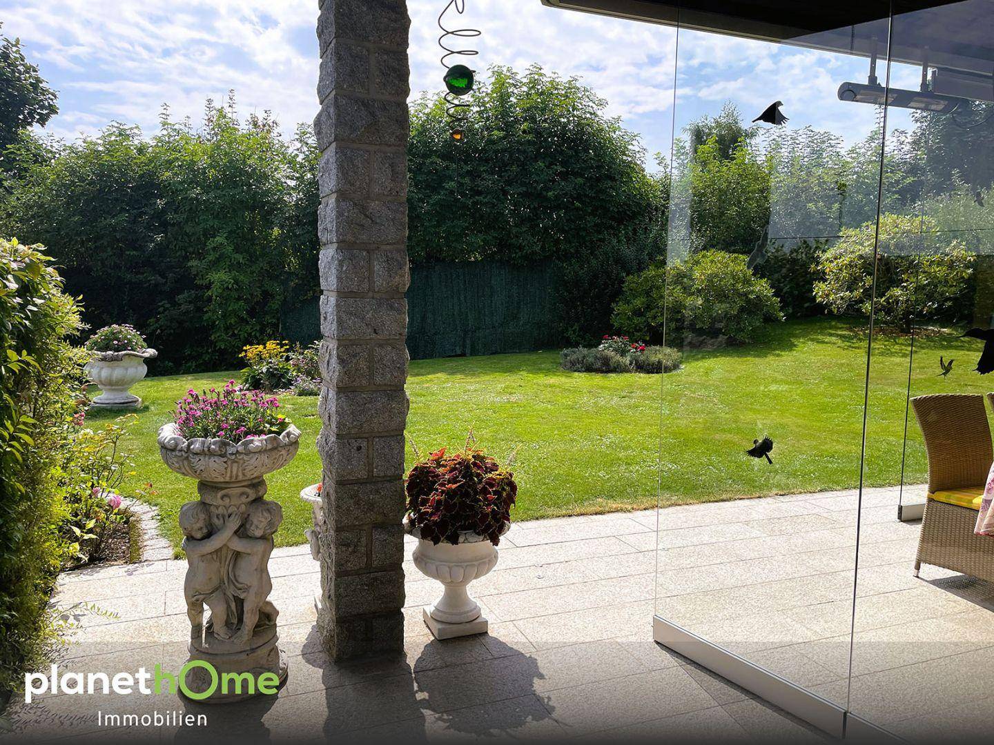 Blick von der verglasten Terrasse in den gepflegten Garten mit Rasenfläche und Hecke.