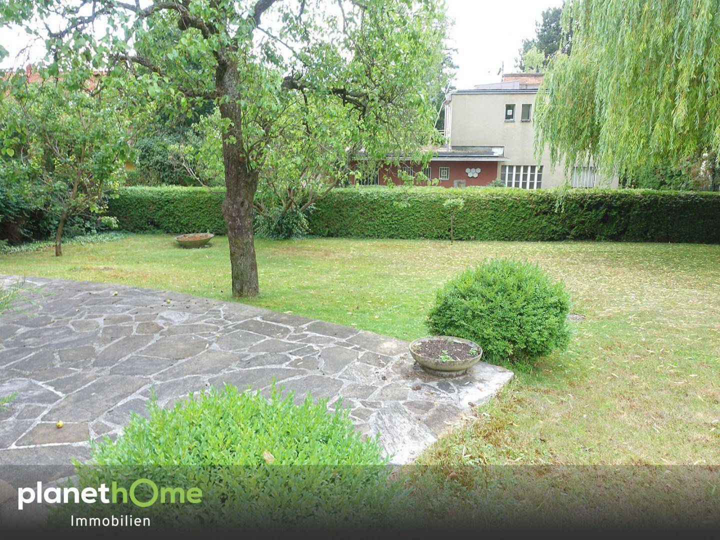 Weitläufiger Garten mit Rasen, Sträuchern und einer Steinplatte, im Hintergrund ist ein Gebäude sichtbar.