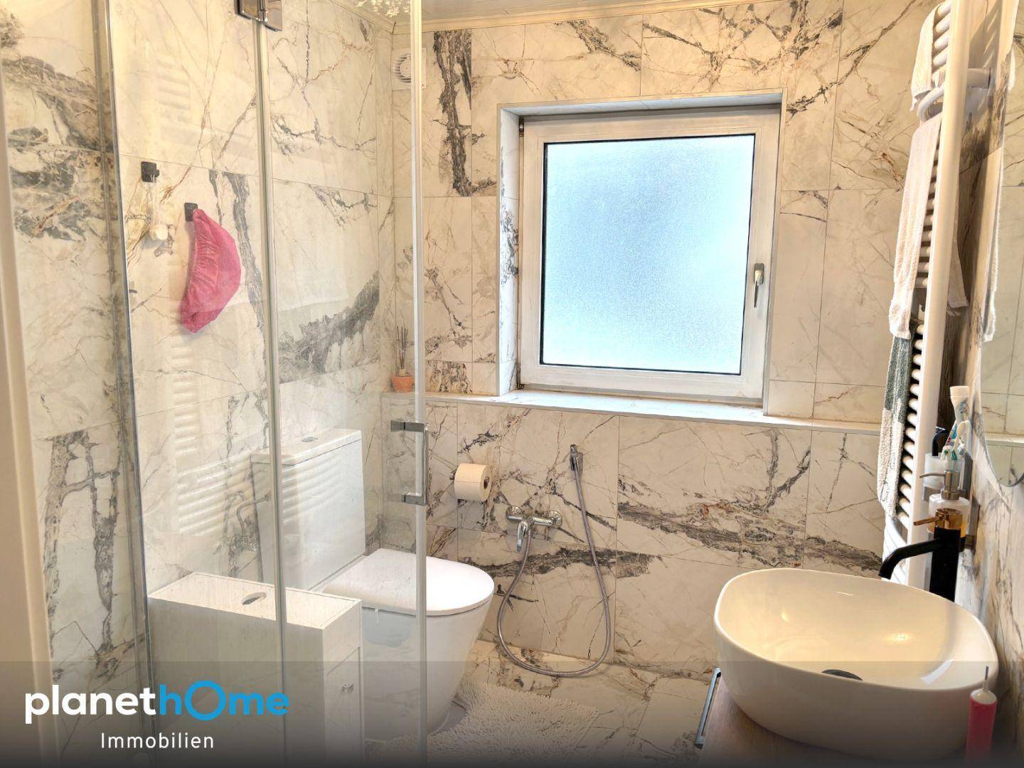 Modernes Badezimmer mit Marmorfliesen, Duschkabine, WC und Fenster für Tageslicht.