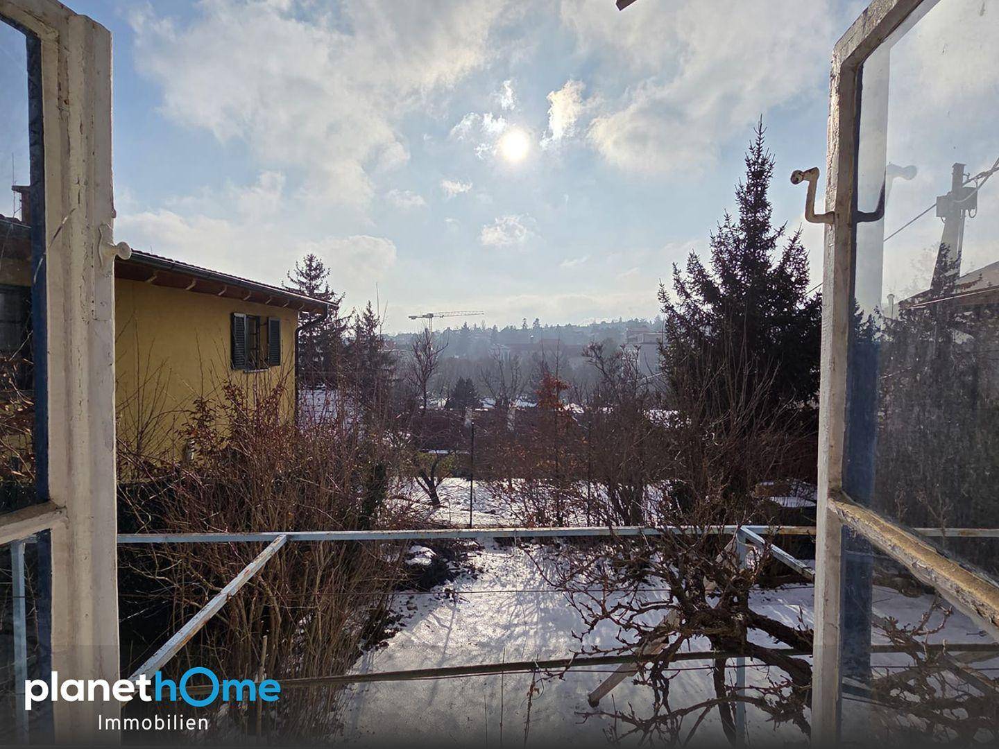 Blick aus einem Fenster auf eine verschneite Landschaft mit Bäumen und entfernten Gebäuden unter sonnigem Himmel.