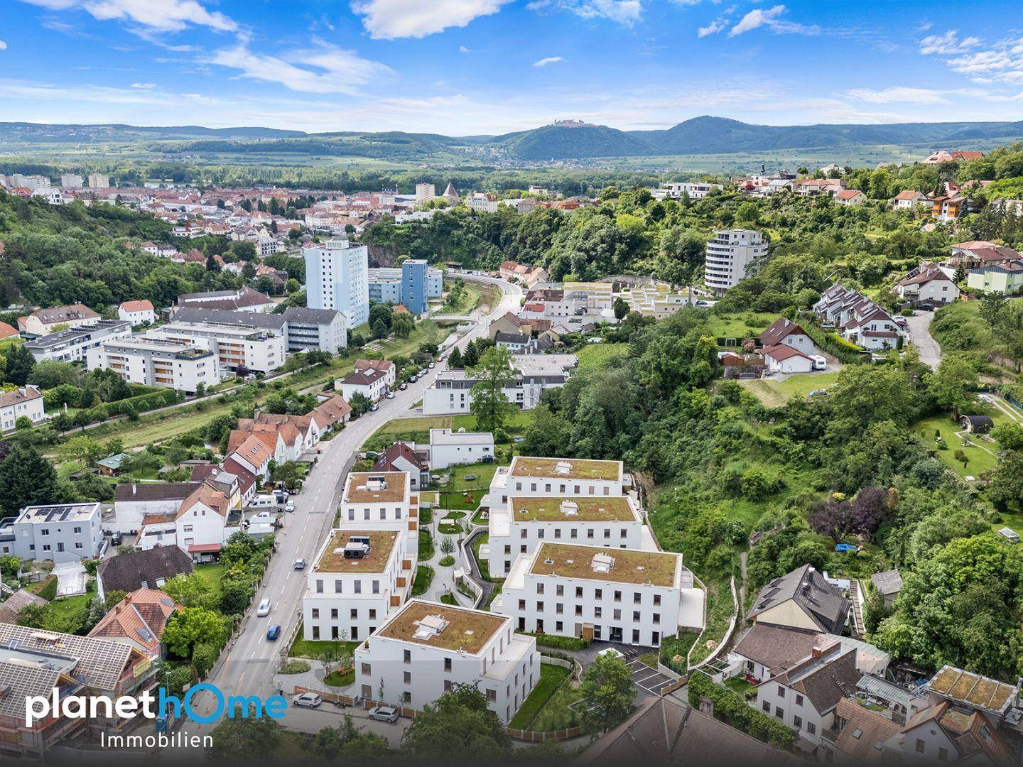 Luftaufnahme einer modernen Wohnsiedlung eingebettet in eine grüne Hügellandschaft mit Stadtblick.