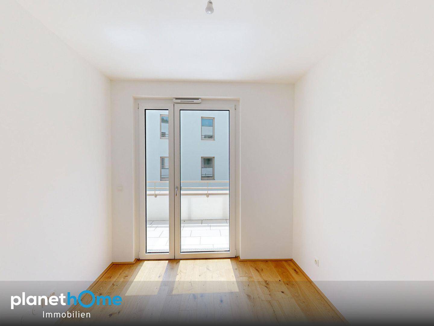 Geräumiges Zimmer mit Holzboden und großen Glastüren, die viel Tageslicht hereinlassen.