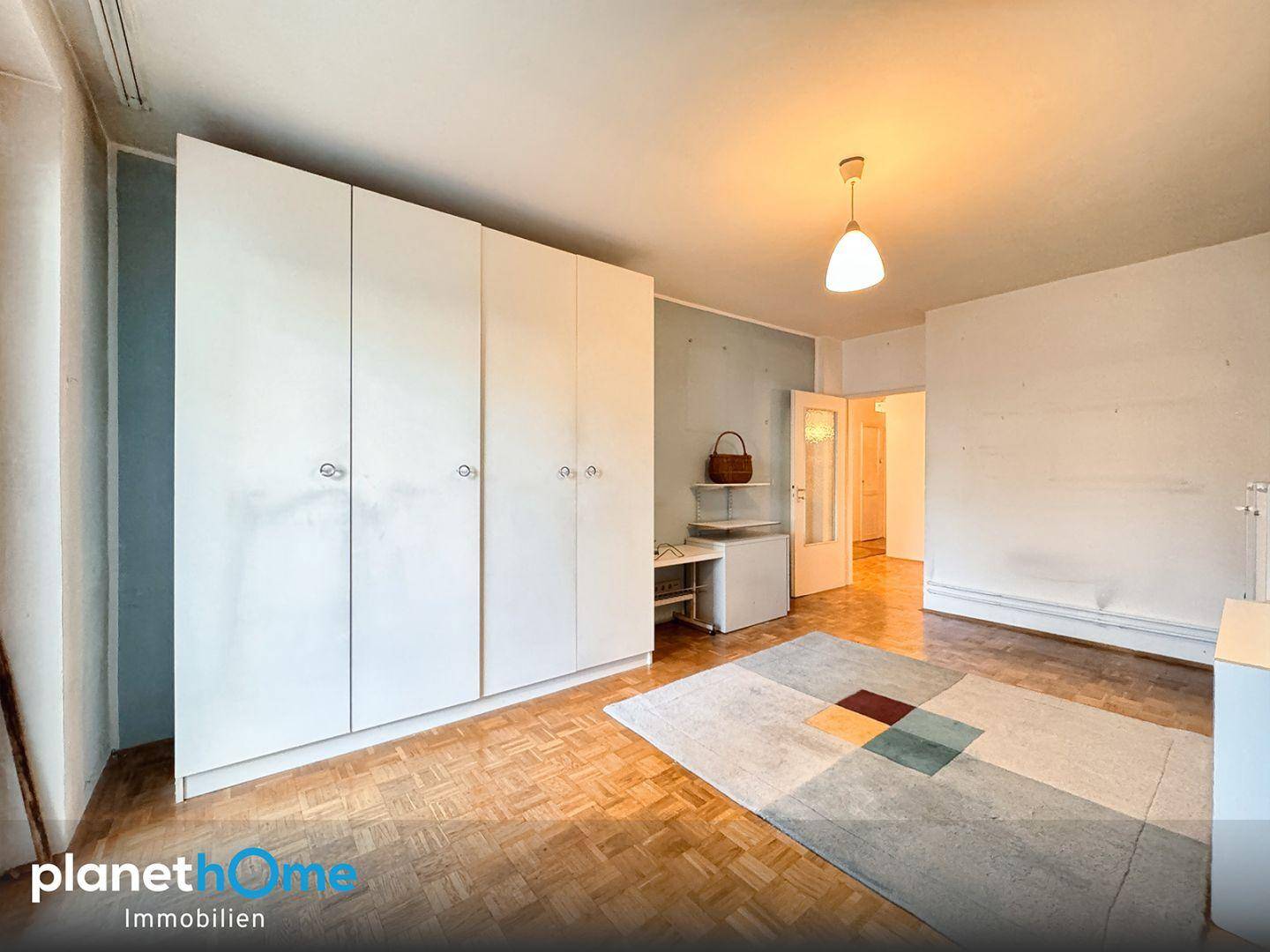 Schlafzimmer mit Parkettboden, großem weißen Kleiderschrank und einem Teppich mit geometrischem Muster.