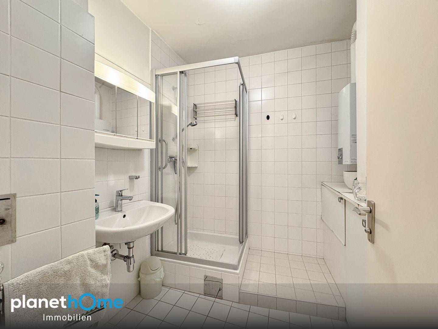 Helles Badezimmer mit Duschkabine, Waschbecken und weißen Fliesen an Wänden und Boden.