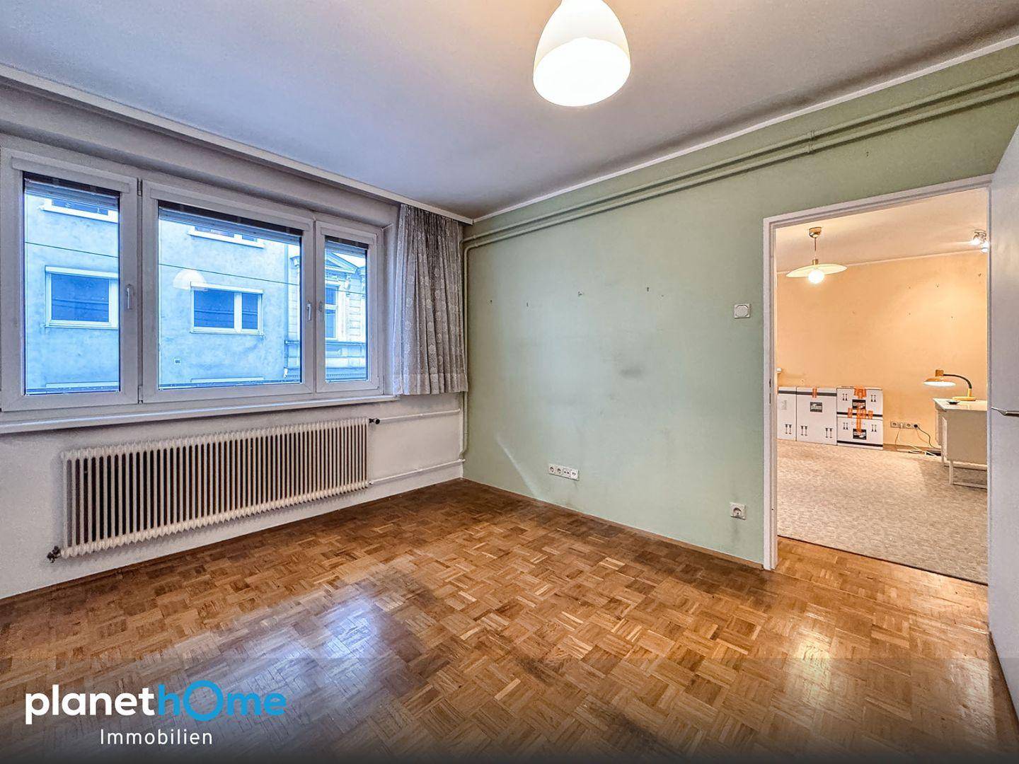Leeres Zimmer mit Parkettboden, grünen Wänden und einem Fenster mit Blick auf die Straße.