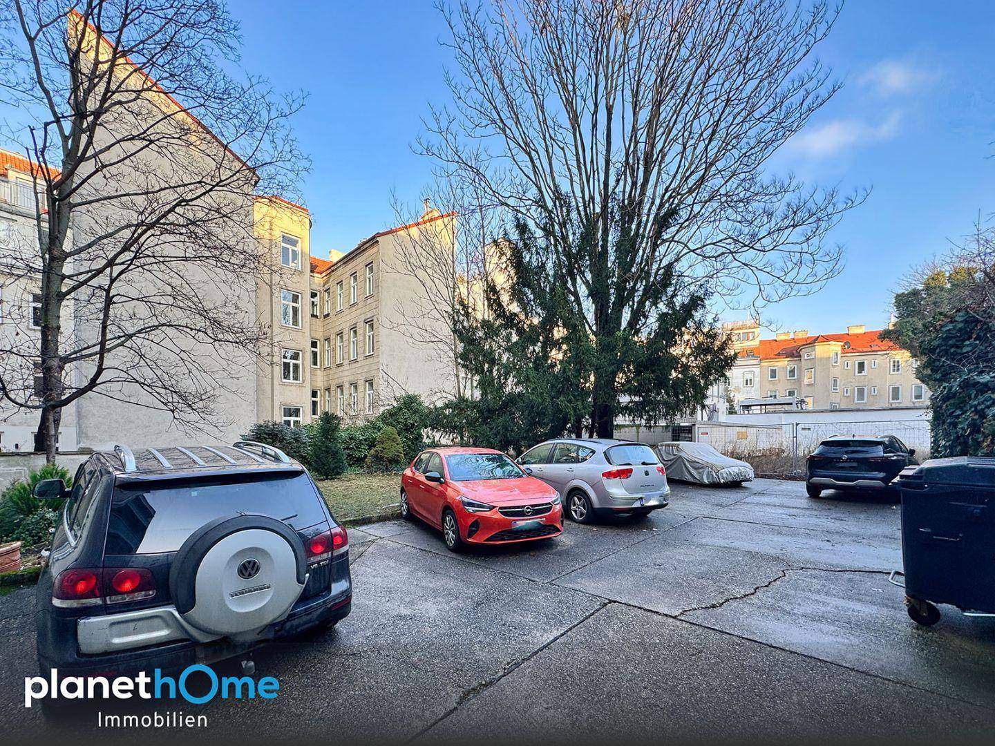 Außenparkplatz mit mehreren Autos und älteren Gebäuden im Hintergrund unter blauem Himmel.