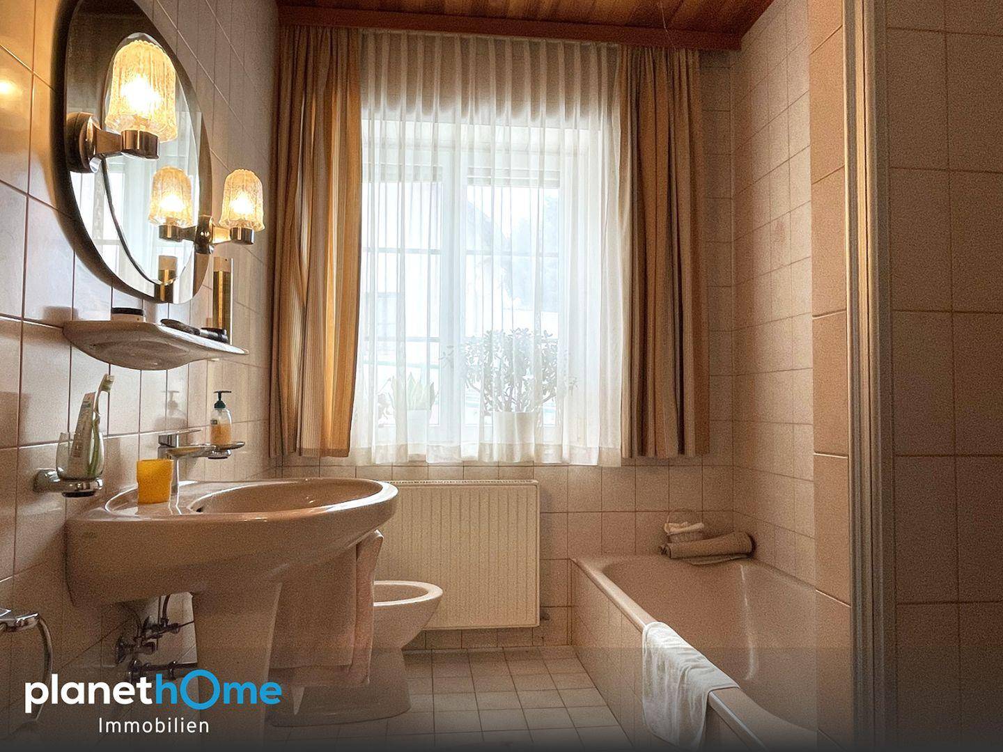 Helles Badezimmer mit Badewanne, Waschbecken und Fenster in beigen Fliesen.