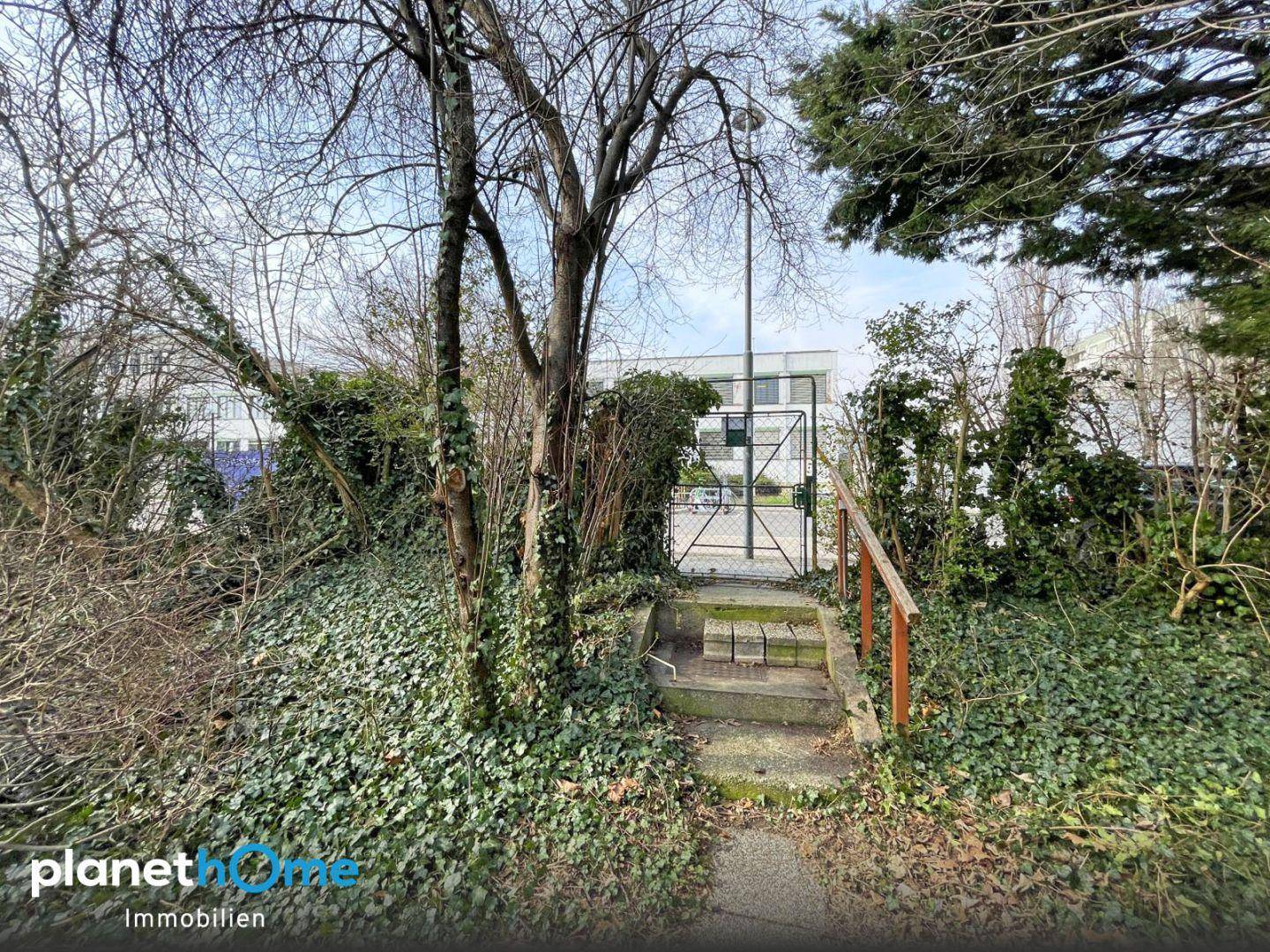 Eingang zum Grundstück mit Stufen, einem Tor und üppiger Vegetation, Gebäude im Hintergrund.