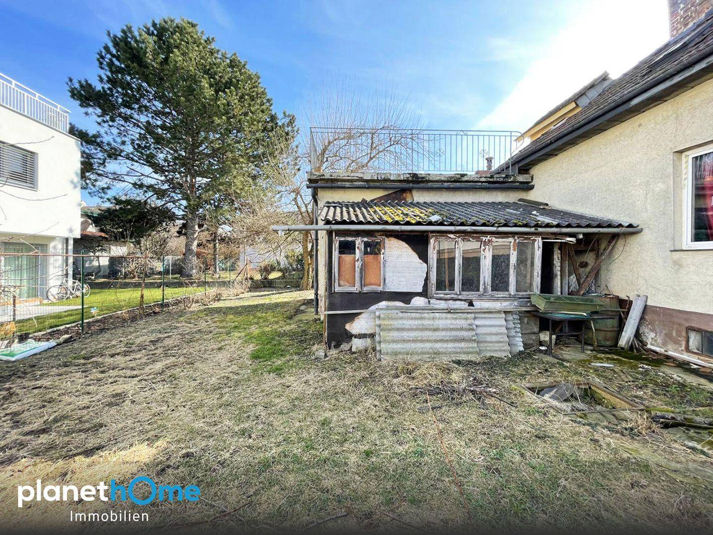 Rückansicht des Hauses mit verfallenem Anbau und modernen Gebäuden im Hintergrund unter blauem Himmel.