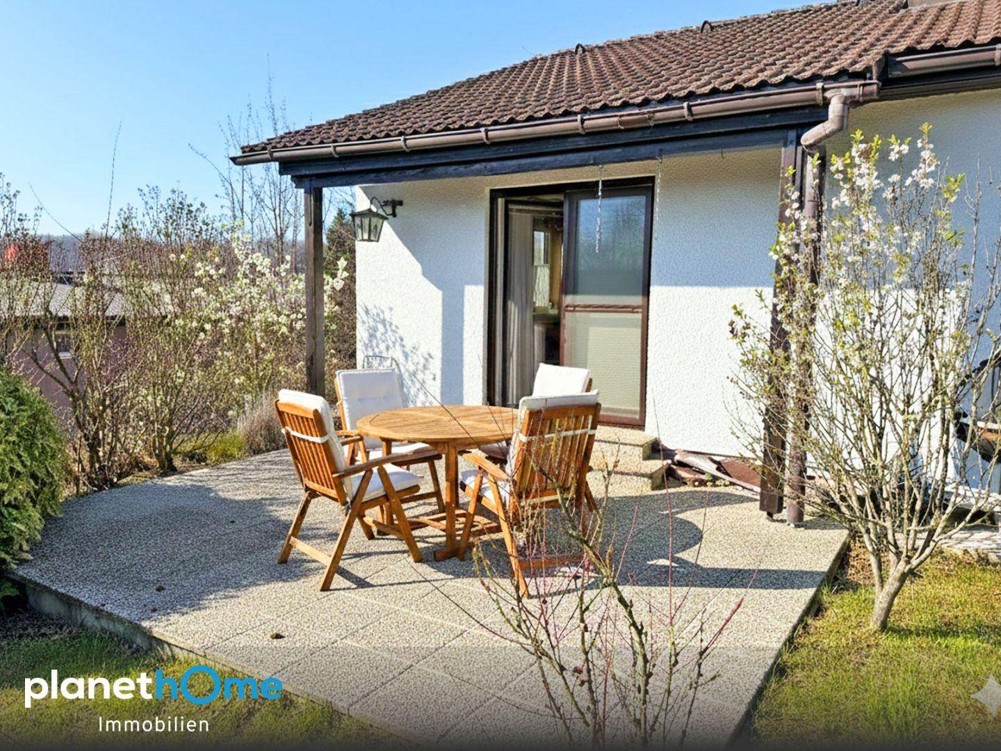 Gemütliche Terrasse mit Holztisch und Stühlen, umgeben von blühenden Sträuchern.