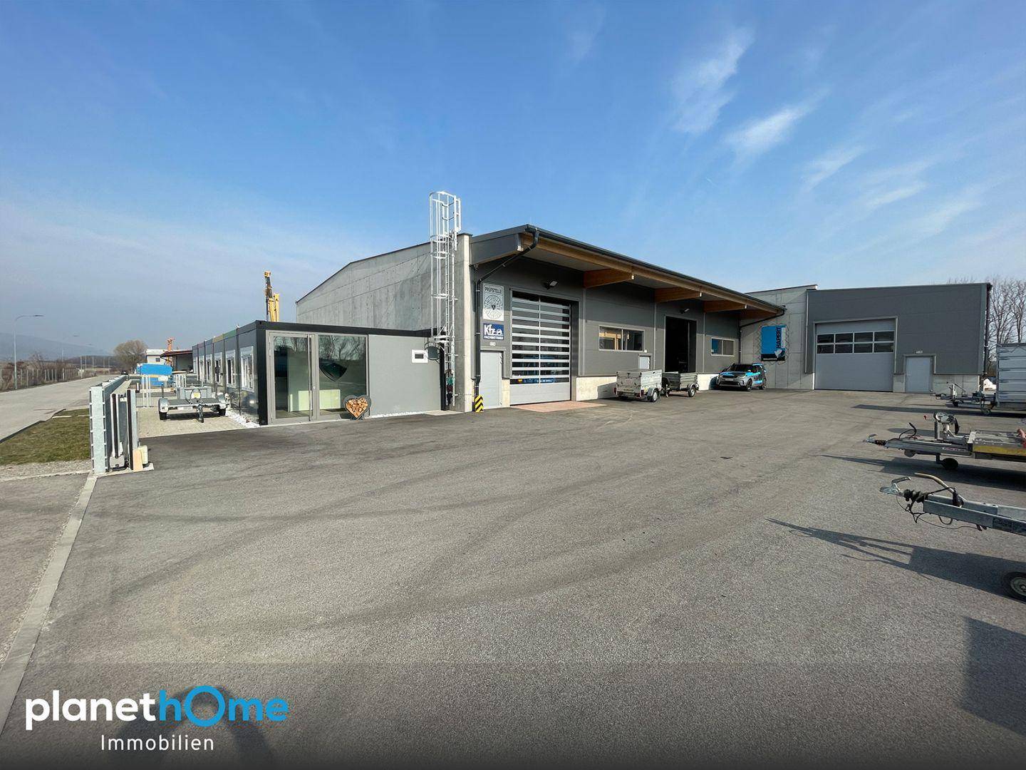 Weitwinkelansicht eines modernen Gewerbegebäudes mit Rolltor und angrenzendem Bürocontainer unter blauem Himmel.