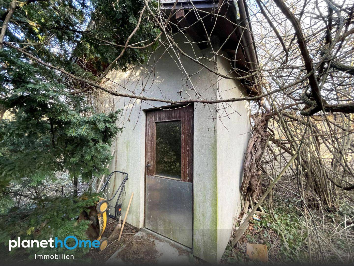 Kleiner Geräteschuppen im Garten, umgeben von Bäumen und Büschen.