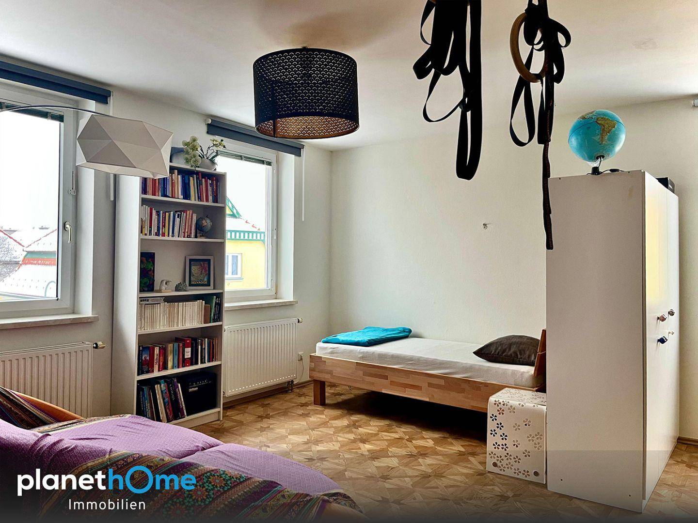 Helles Schlafzimmer mit Einzelbett, Bücherregal und zwei Fenstern.