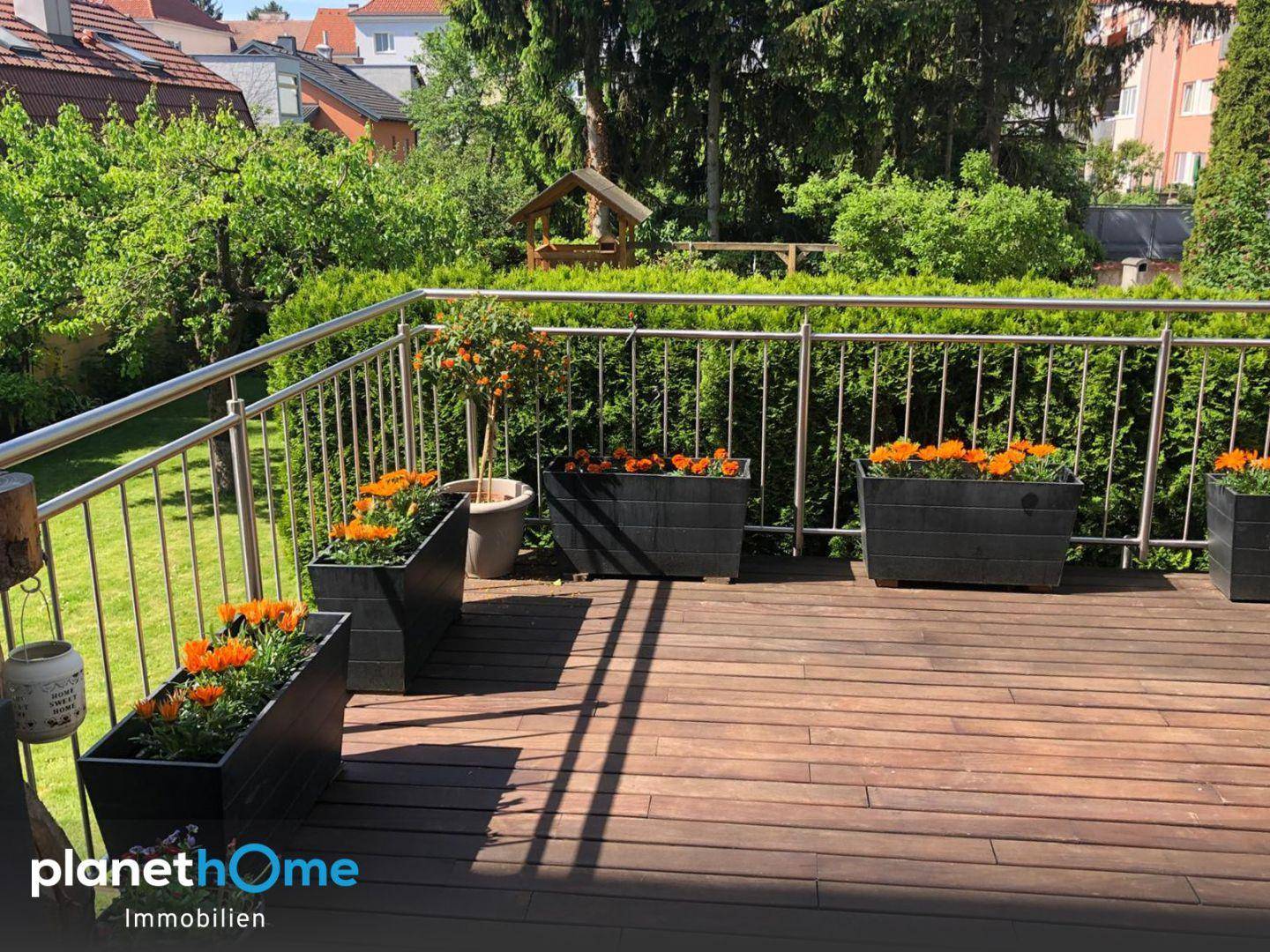 Holzterrasse mit Metallgeländer und Blumenkästen, umgeben von viel Grün.