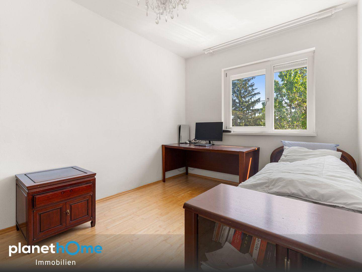 Helles Zimmer mit Holzboden, Schreibtisch und einem Fenster mit Blick ins Grüne.