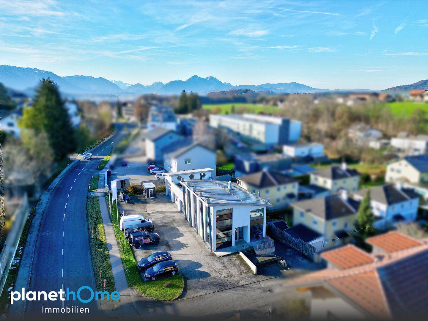 Weitläufige Luftaufnahme des Geschäftsgebäudes mit Straße und beeindruckendem Alpenpanorama im Hintergrund.