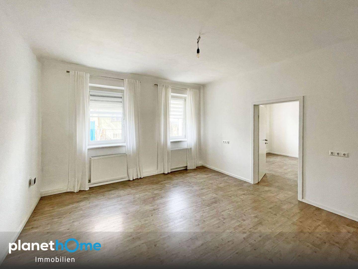 Helles Wohnzimmer mit Laminatboden, weißen Wänden und zwei Fenstern.