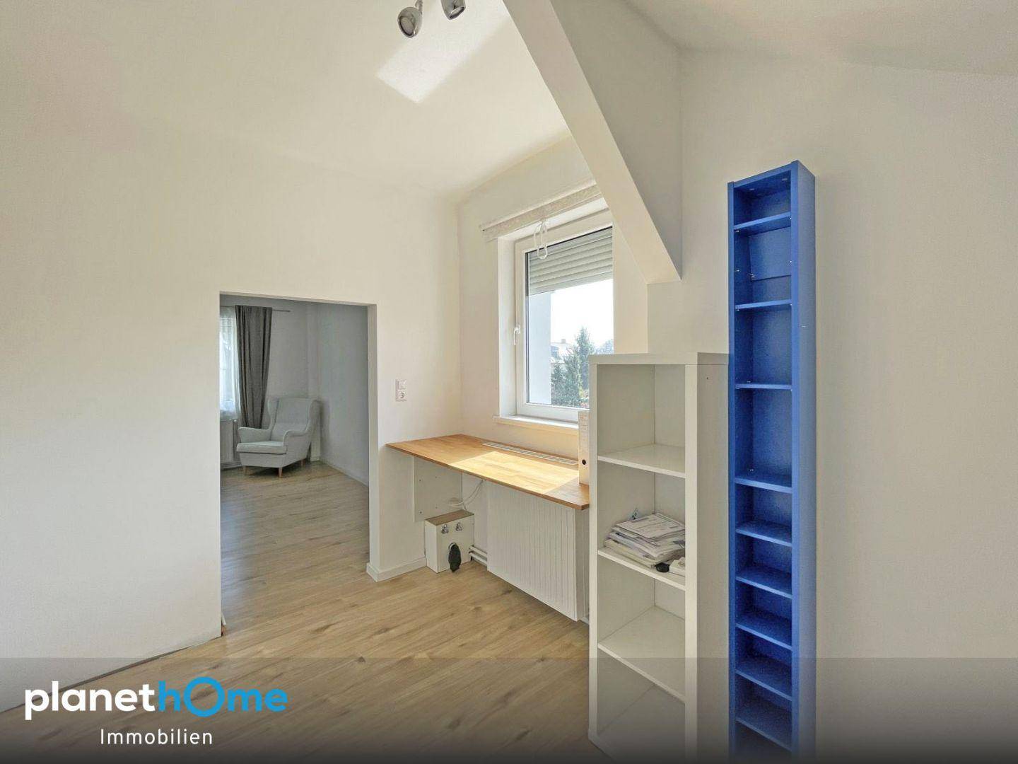 Dachzimmer mit Schreibtisch, Regal und Zugang zu einem weiteren Raum.