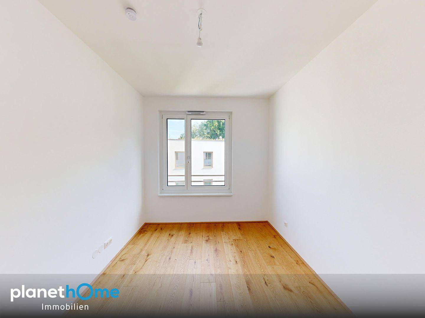 Leeres, helles Zimmer mit Holzfußboden und einem Fenster, das viel Tageslicht hereinlässt.