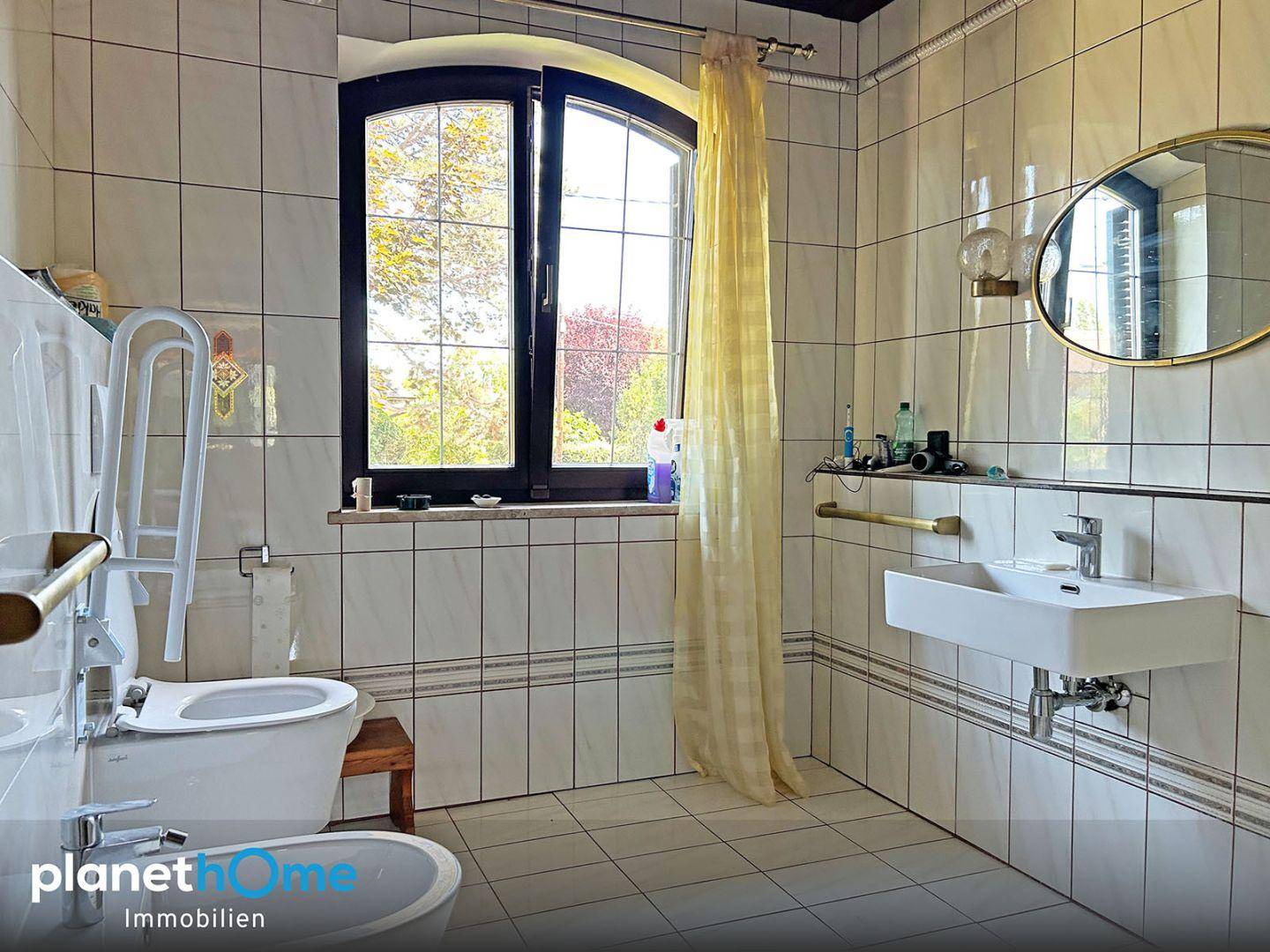 Gefliestes Badezimmer mit WC, Bidet, Waschbecken und Fenster, ausgestattet mit Haltegriffen.