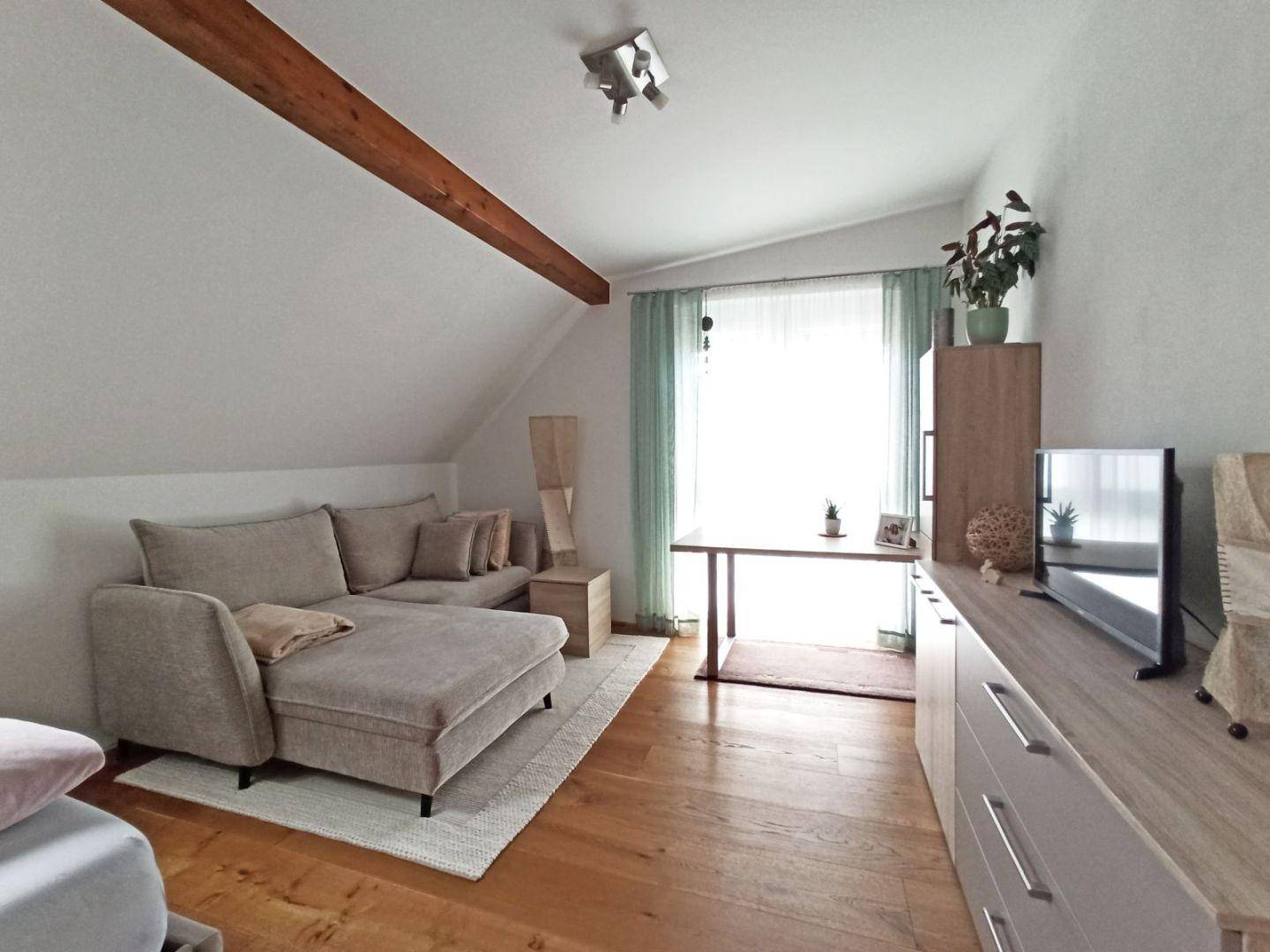 Helles Zimmer mit Dachschräge, Holzbalken, Sofa und Arbeitsbereich am Fenster.