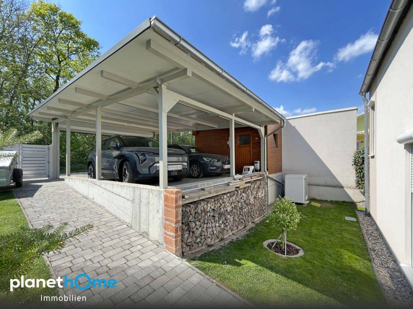 Carport-Bereich mit Holzstapel und Zugang zum Haus, umgeben von Grünflächen.