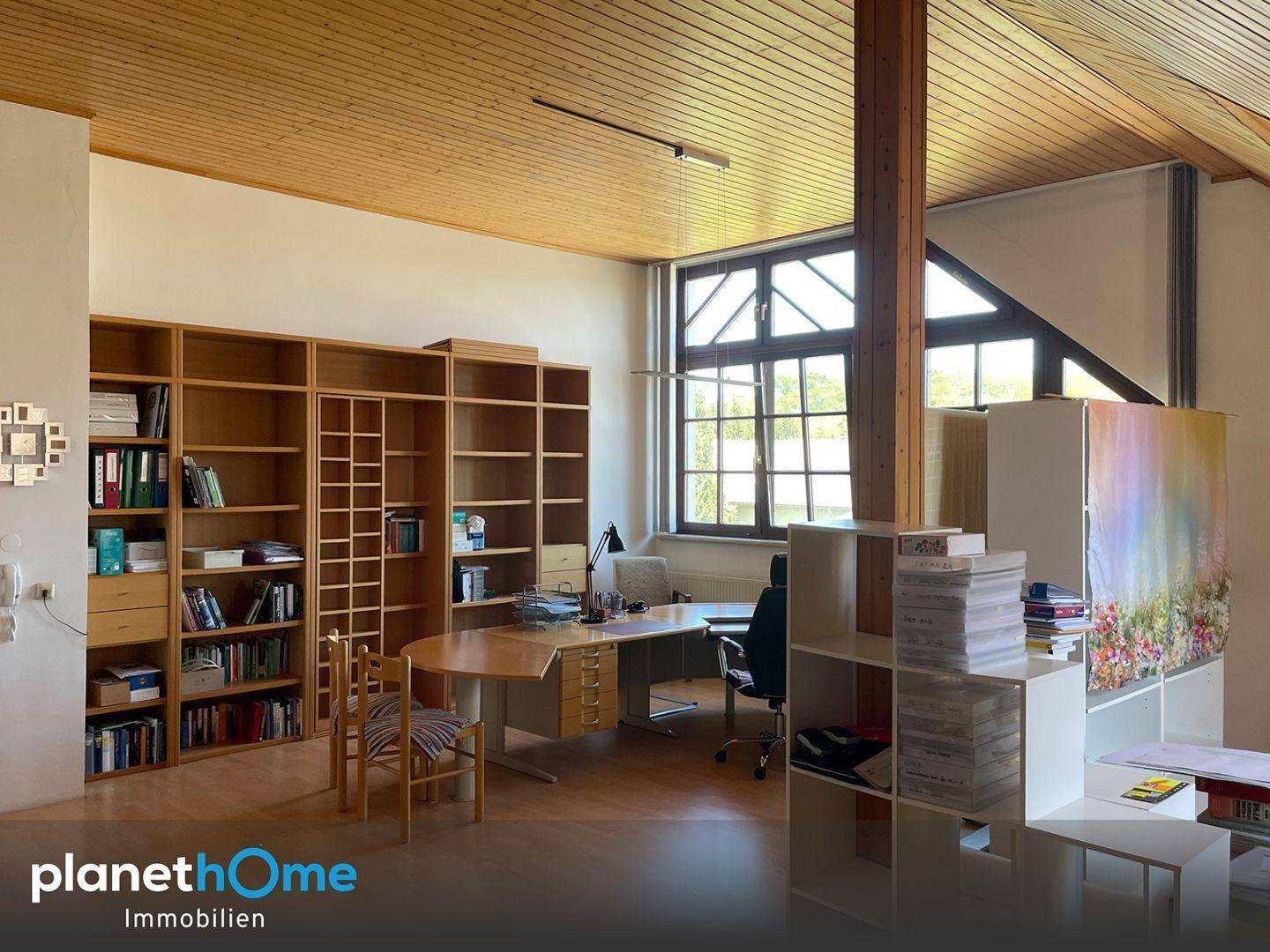 Geräumiges Büro mit Holzdecken, großen Fenstern und umfangreichen Bücherregalen in hellem Ambiente.