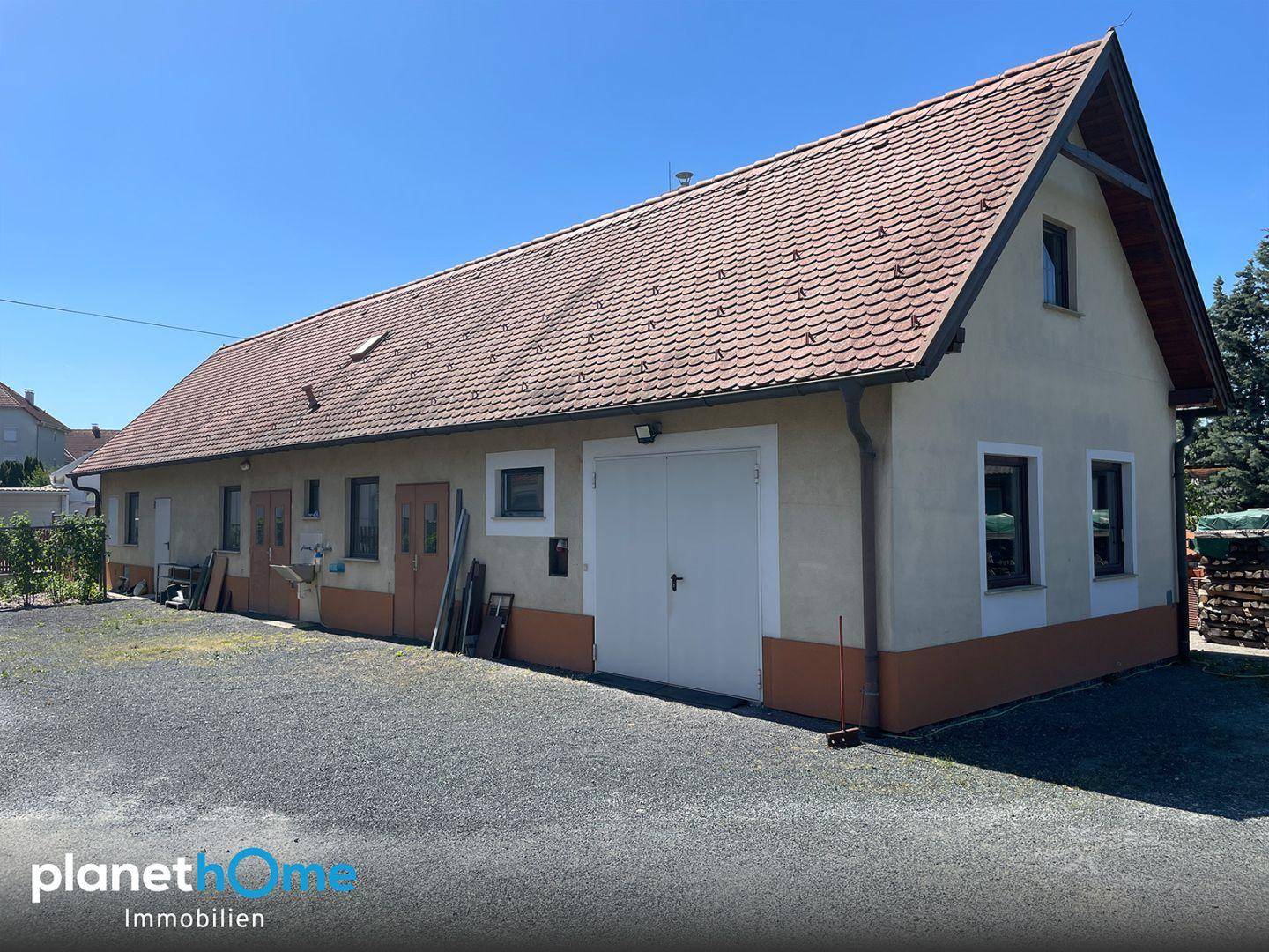 Langes Gebäude mit Satteldach, Garagentor und großem Vorplatz unter strahlendem Sonnenschein.
