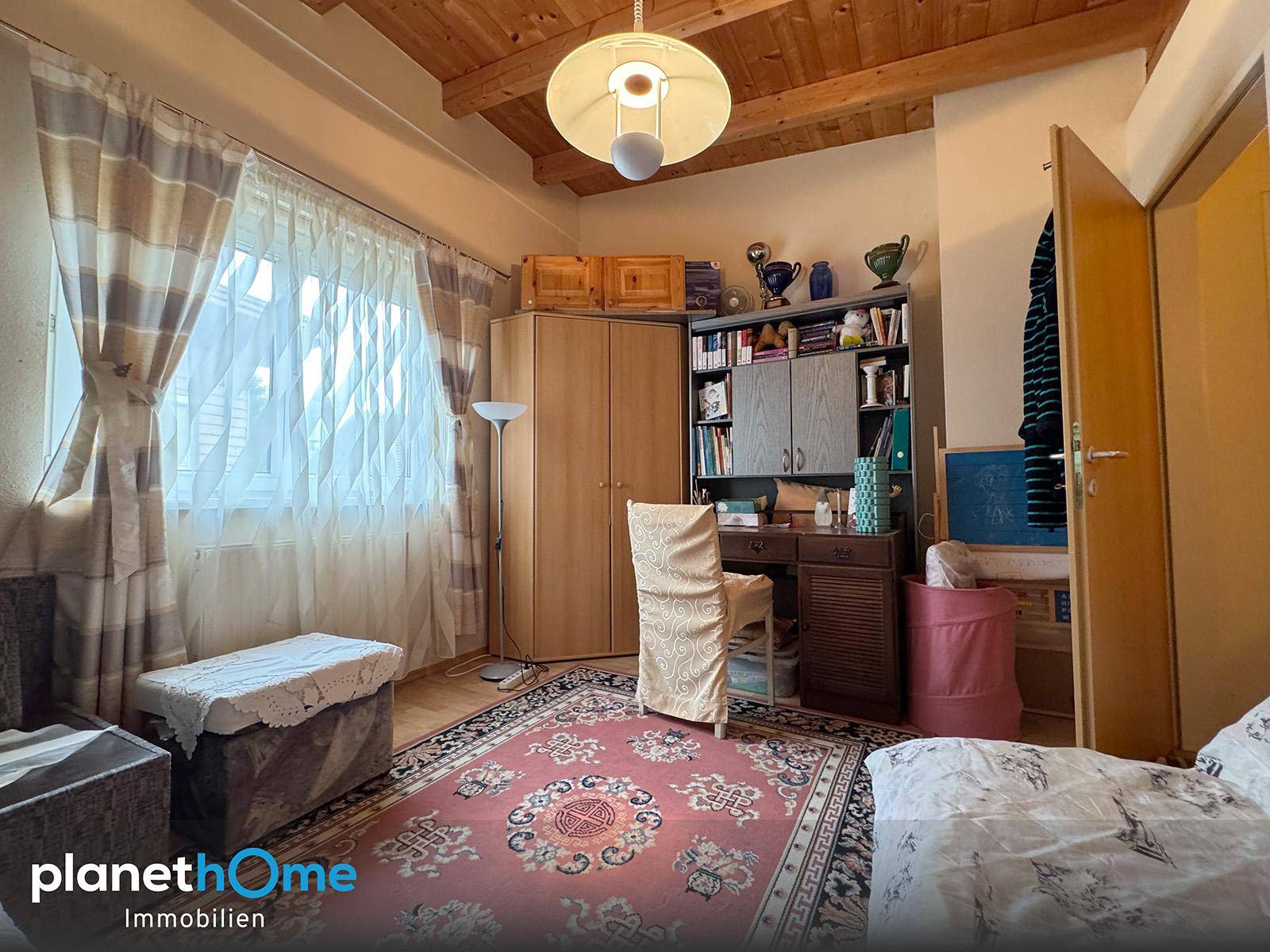 Schlafzimmer mit Holzbalkendecke, Teppichboden und Schreibtischbereich am Fenster.