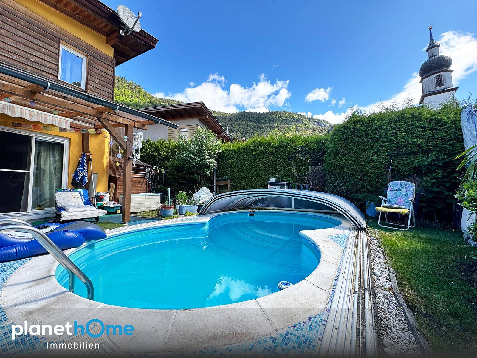 Garten mit Pool und Blick auf die Kirche und die umliegende Berglandschaft unter blauem Himmel.