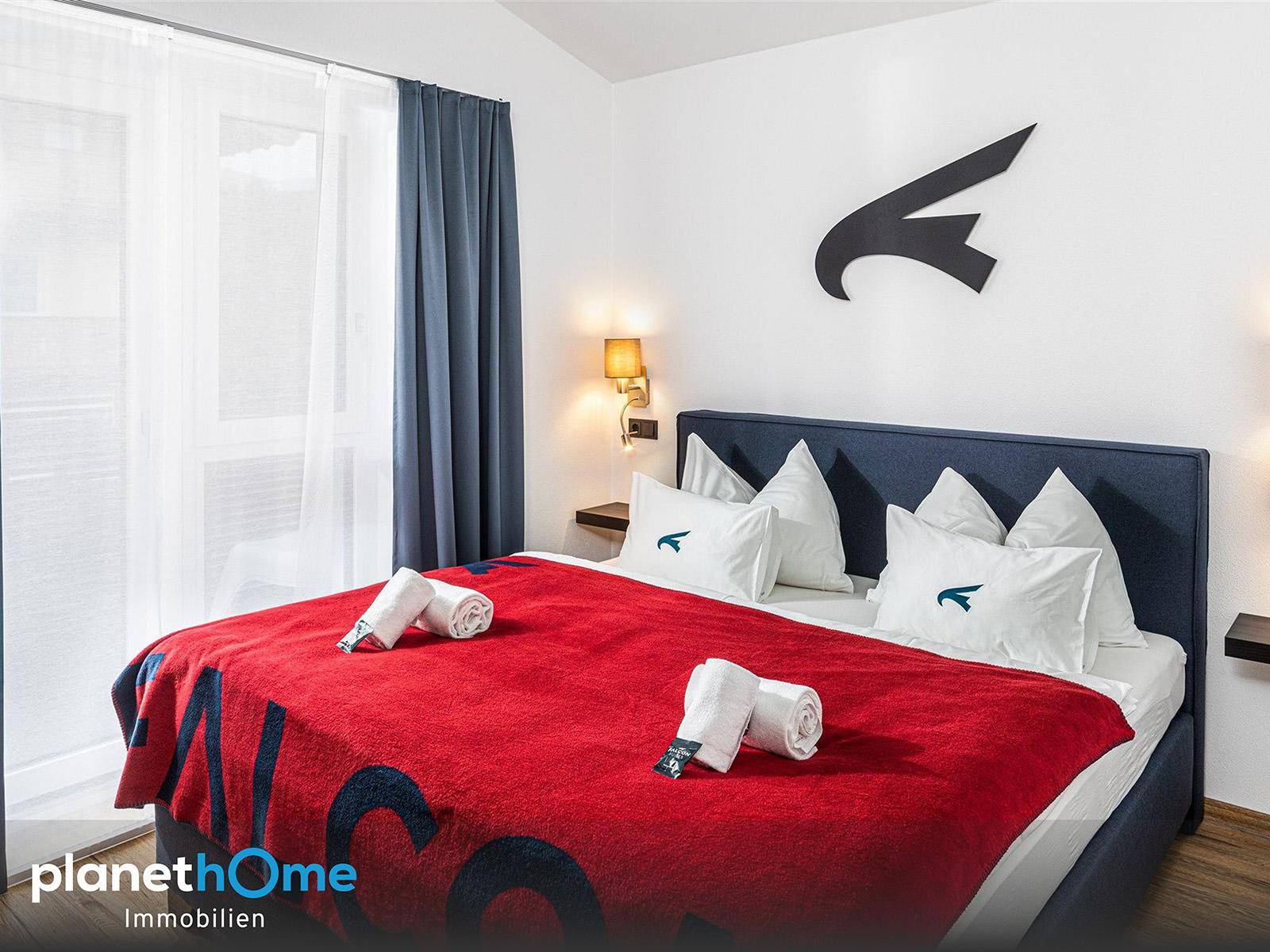 Gemütliches Schlafzimmer mit großem Doppelbett, roter Decke und blauen Vorhängen, hell durch das Fenster.