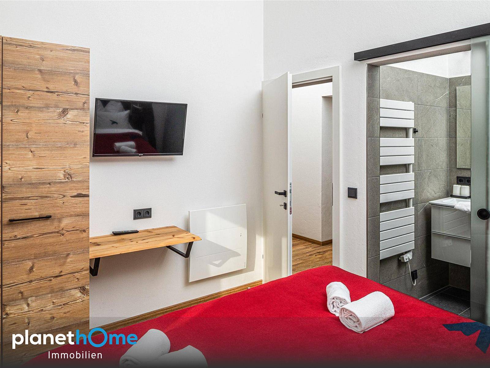 Schlafzimmer mit Holzkleiderschrank und Wandfernseher, angrenzend an ein Badezimmer mit Handtuchheizkörper.