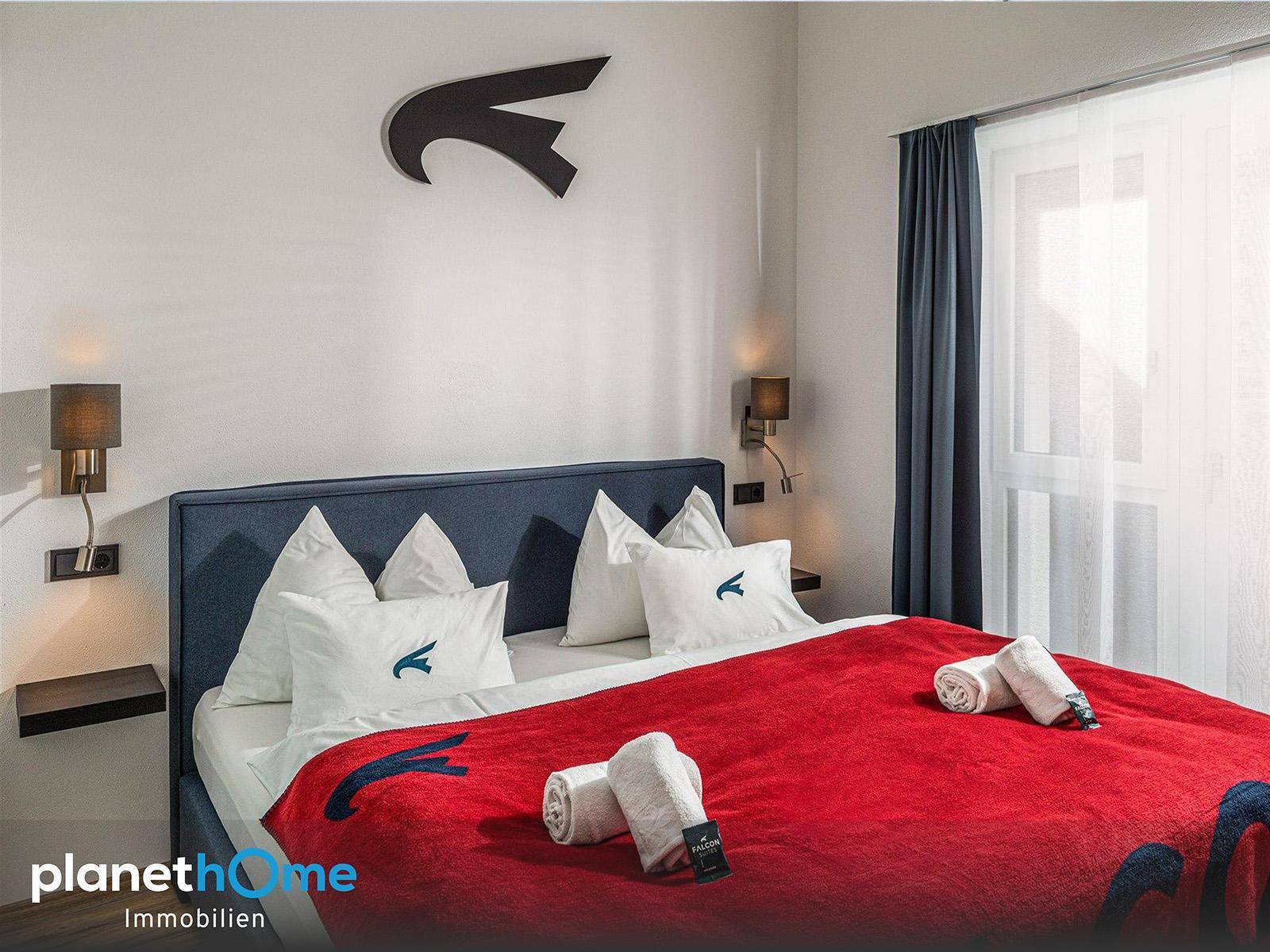 Helles Schlafzimmer mit großem Doppelbett, roter Decke und blauen Vorhängen, mit viel Tageslicht.