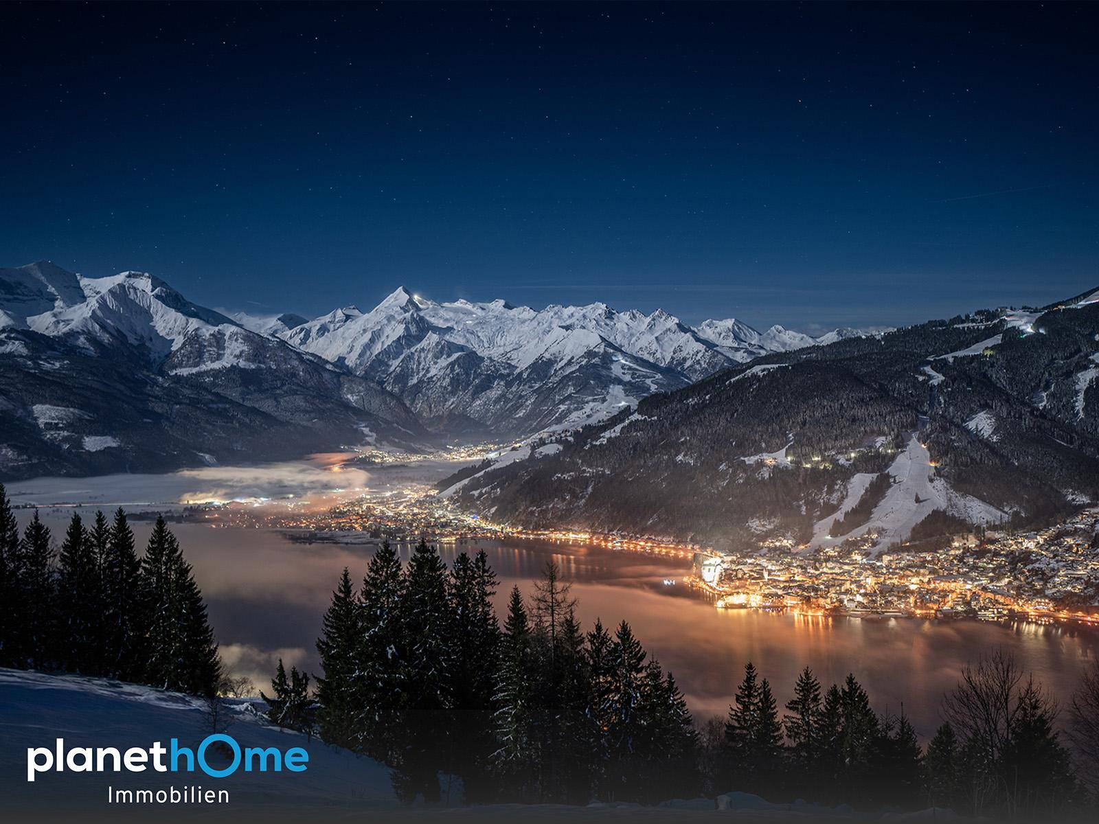 Beeindruckender Nachtblick auf einen See und beleuchtete Stadt, umgeben von schneebedeckten Bergen unter klarem Himmel.
