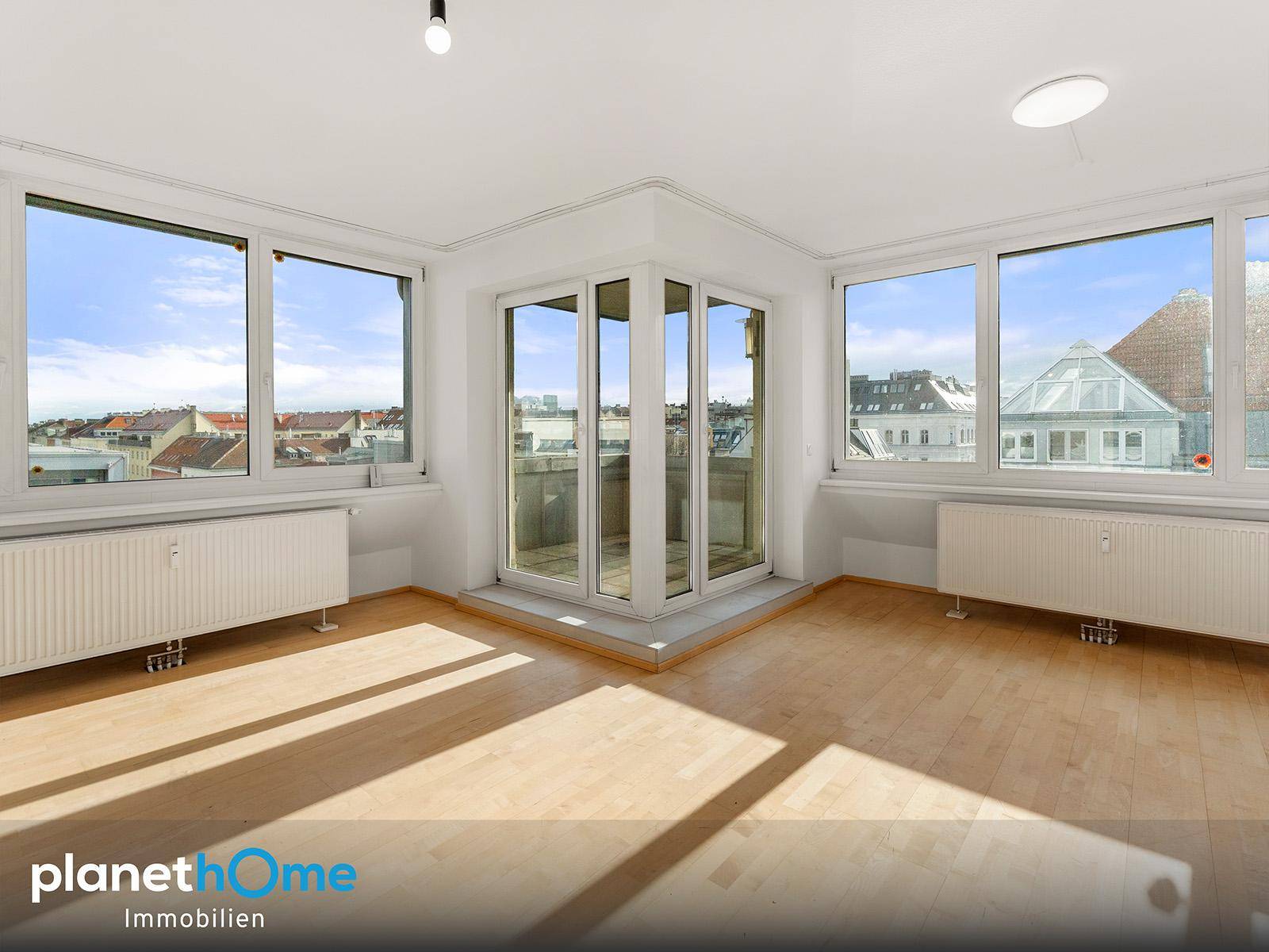 Heller, leerer Raum mit großen Fenstern, Holzboden und Zugang zu einem Eckbalkon mit Stadtblick.