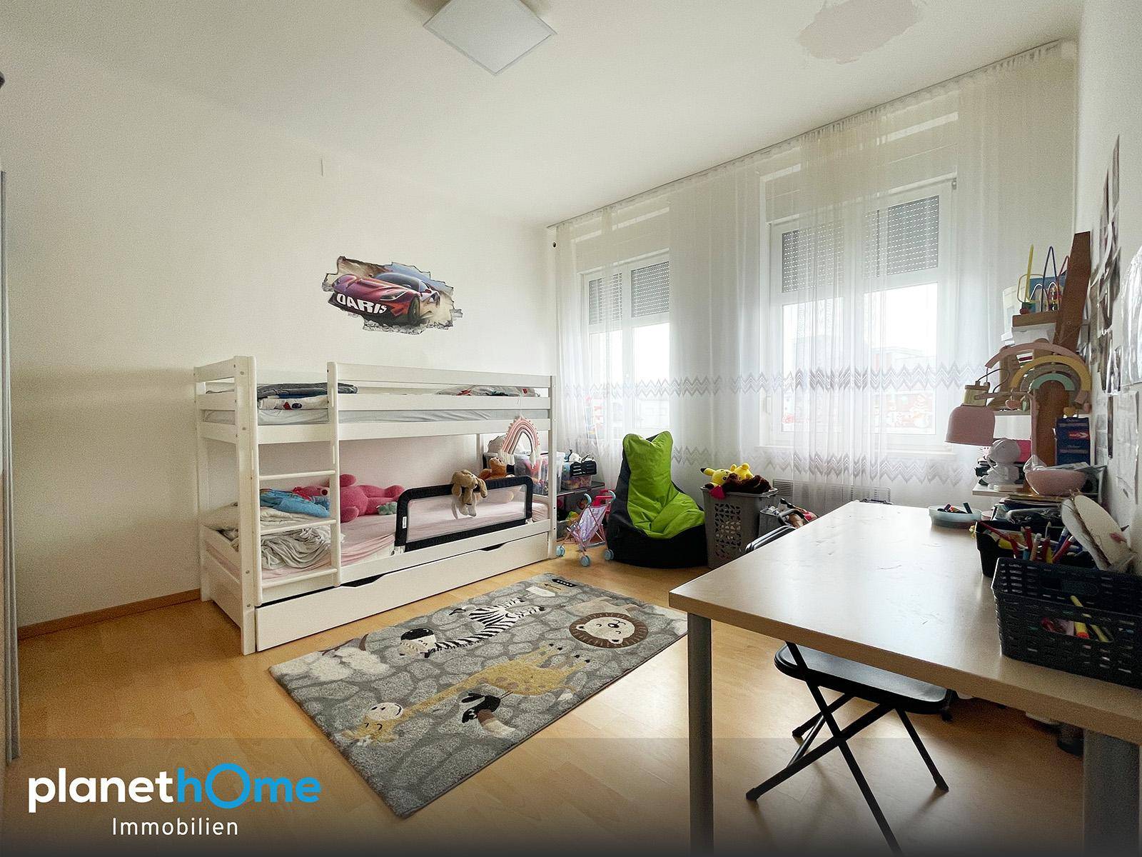 Kinderzimmer mit Etagenbett, Schreibtisch und Teppich, hell durch große Fenster.