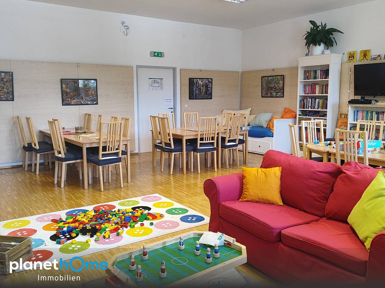Großer Gemeinschaftsraum mit Spielbereich, Esstischen und einem roten Sofa für Entspannung.