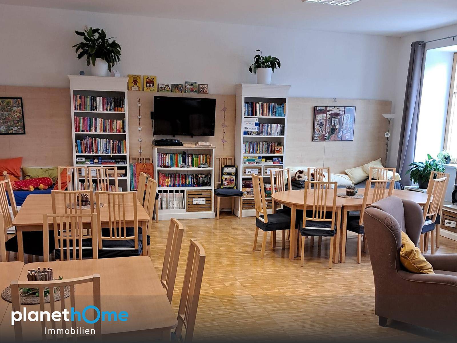 Geräumiger Gemeinschaftsraum mit mehreren Esstischen, Bücherregalen und einem gemütlichen Sessel.