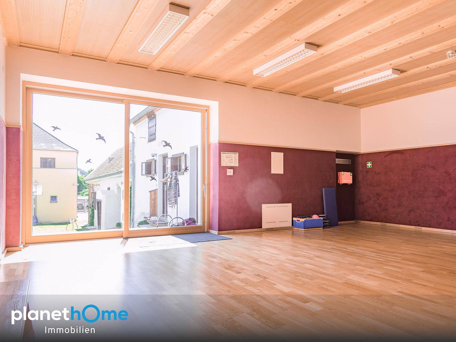 Großer Mehrzweckraum mit Holzboden, Holzdecke und einer breiten Glasfront zur Außenseite.