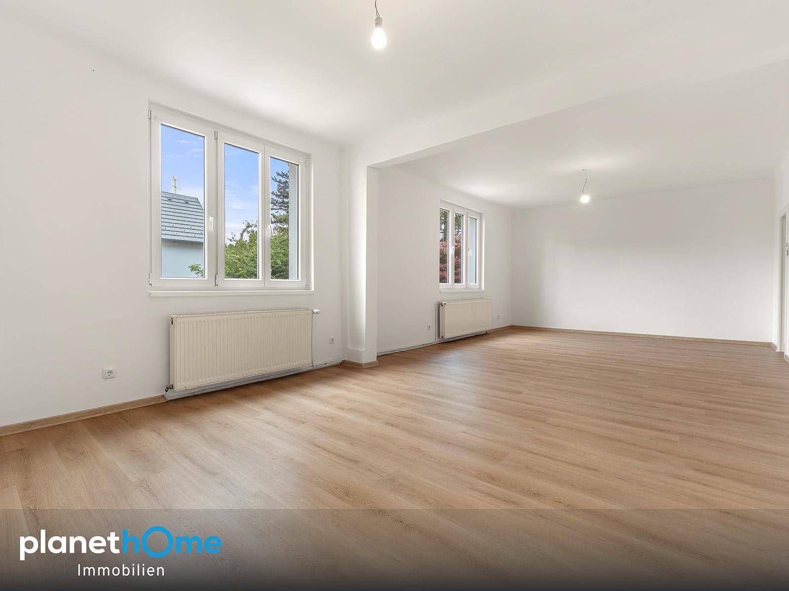 Helles Wohnzimmer mit Laminatboden und mehreren Fenstern, die viel Tageslicht hereinlassen.