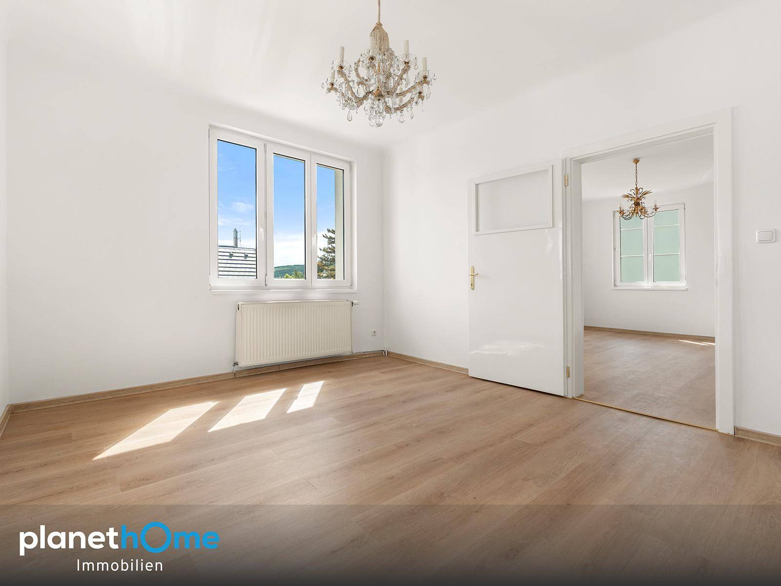 Helles Wohnzimmer mit Laminatboden, großen Fenstern und einem Kronleuchter.