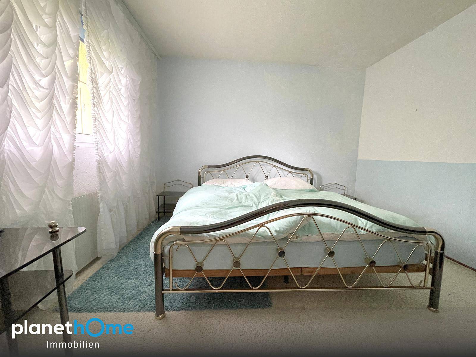 Schlafzimmer mit Metallbett, hellblauen Wänden und Vorhängen, die viel Licht hereinlassen.