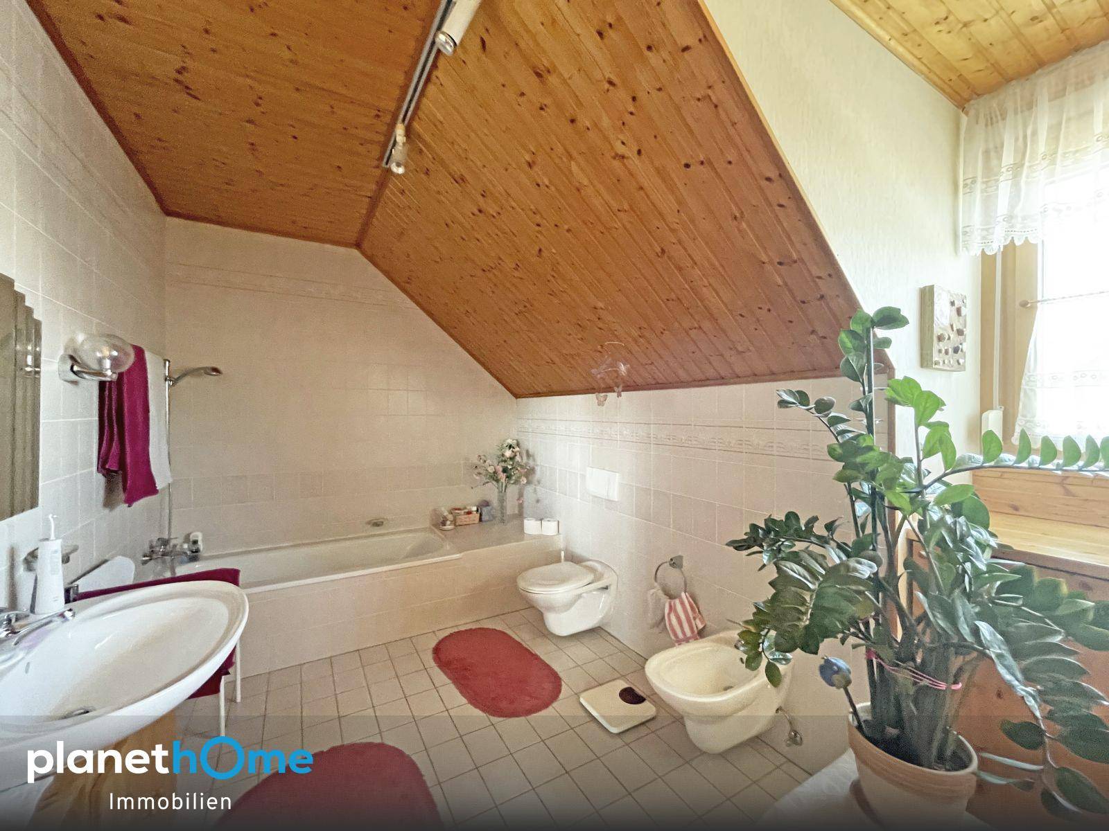 Badezimmer im Dachgeschoss mit Badewanne, WC, Bidet und Holzdecke, hell gefliest.
